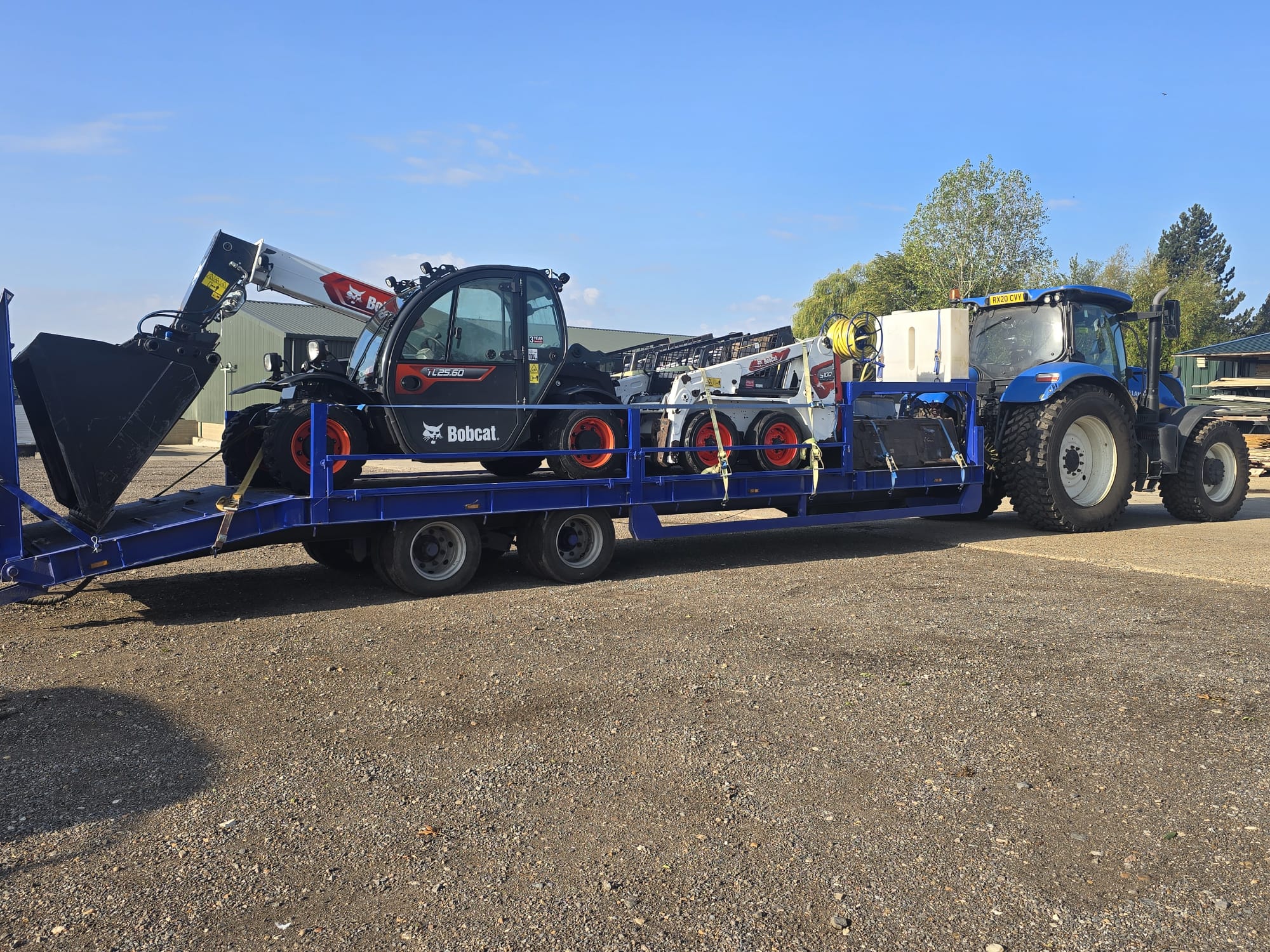 New Holland tractor pulling a low-loader carrying a Bobcat telehandler and S100 skid steer