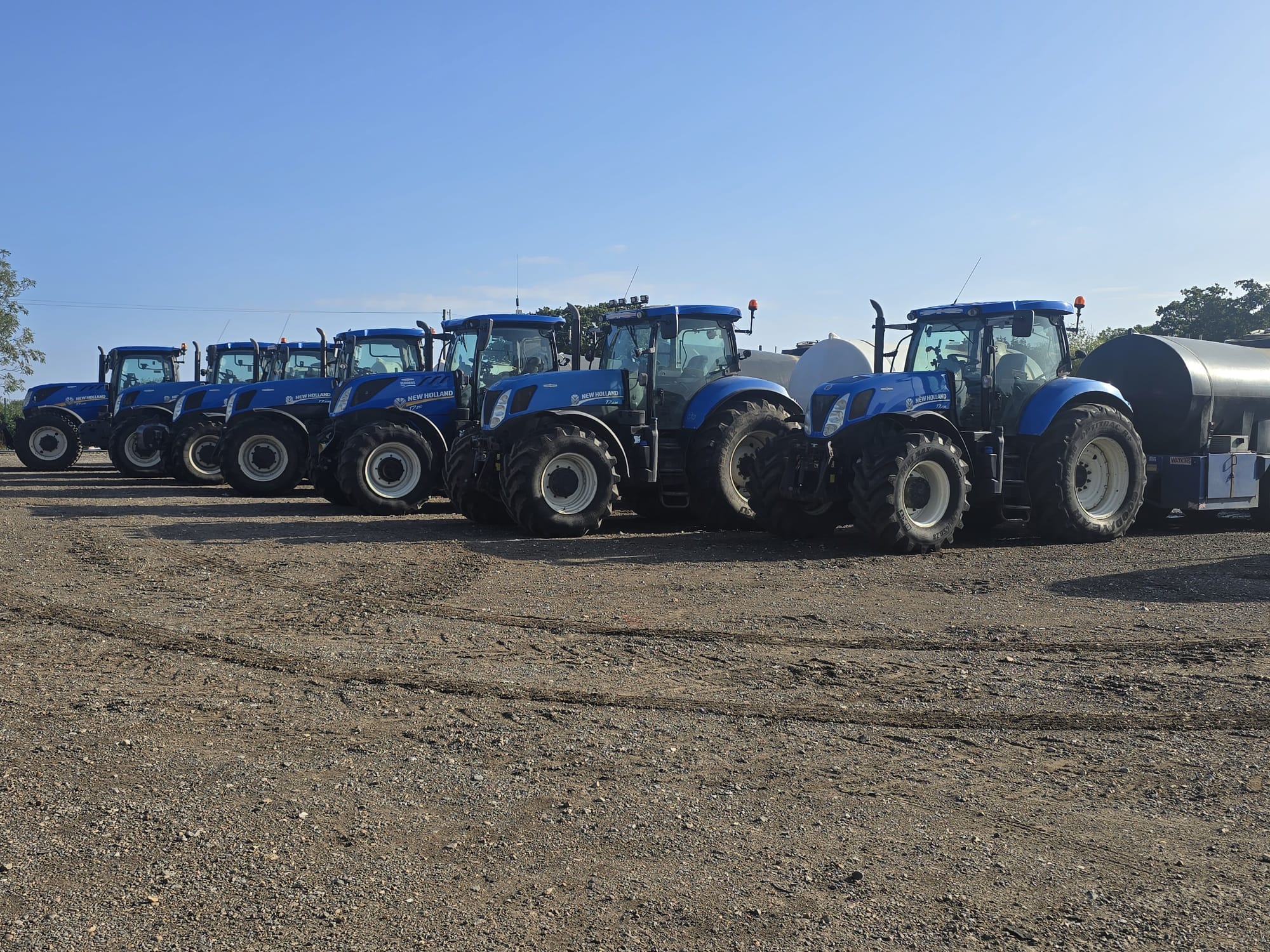 Full line-up of New Holland tractors with bowsers on a sunny day